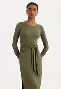 Vestido midi em verde oliva, com textura ligeiramente nervurada e mangas de três quartos, apresentando um detalhe de cintura atada, fendas laterais e bolsos. Textura suave.