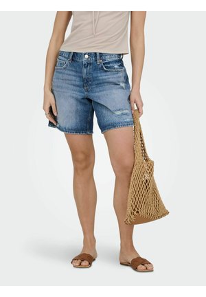 Femme portant un haut beige clair, un short en denim bleu délavé, des sandales plates marron, tenant un sac fourre-tout tressé camel à son côté.