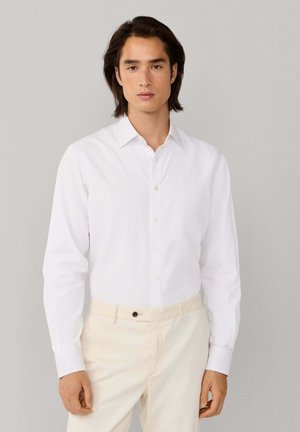 Jeune homme portant une chemise blanche à manches longues boutonnée et un pantalon beige, debout devant un fond gris clair uni.