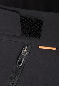 Närbild av en svart dragkedja med tygdragflik, svart kardborrefäste och en liten orange tygdetalj på en mörk textil bakgrund.