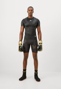Homme athlétique debout, portant un maillot de compression noir, des gants de boxe noirs et dorés, un short noir avec des lettres dorées, et des baskets montantes noires.