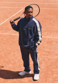 Survêtement marine avec des bandes blanches sur les côtés, col zippé et détail de logo. Le joueur tient une raquette de tennis sur un court en terre battue avec une texture orange.