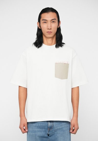 Junger Mann mit langen schwarzen Haaren, der ein weißes, übergroßes T-Shirt mit einer beigen Brusttasche und blauen Jeans trägt, steht vor einem einfarbigen Hintergrund.