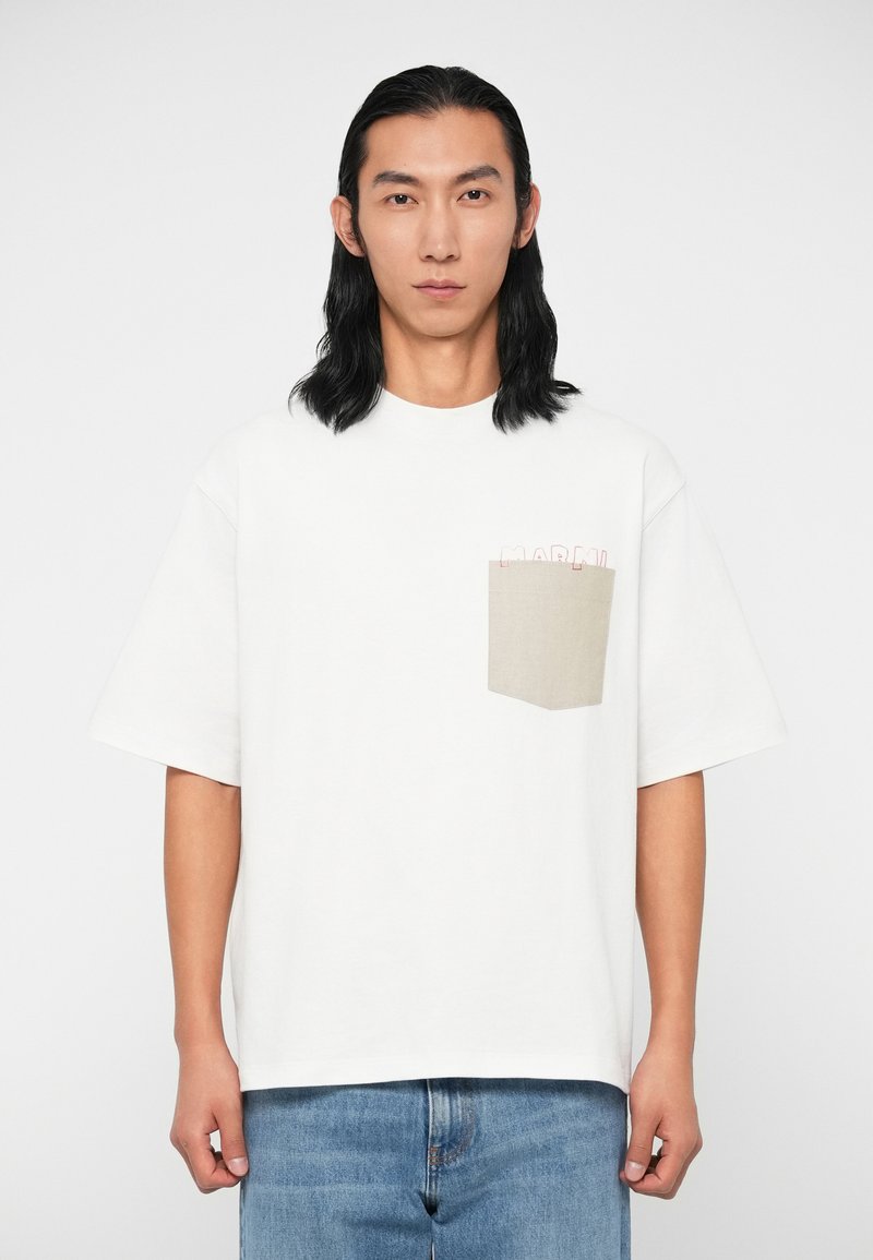Junger Mann mit langen schwarzen Haaren, der ein weißes, übergroßes T-Shirt mit einer beigen Brusttasche und blauen Jeans trägt, steht vor einem einfarbigen Hintergrund.