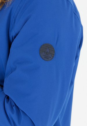 Blå stofjakkeærme med en rund, sort læderpatch med et bjerg- og bølgemotiv. Glat tekstur, afslappet design.