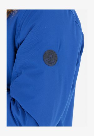 Blå stofjakkeærme med en rund, sort læderpatch med et bjerg- og bølgemotiv. Glat tekstur, afslappet design.