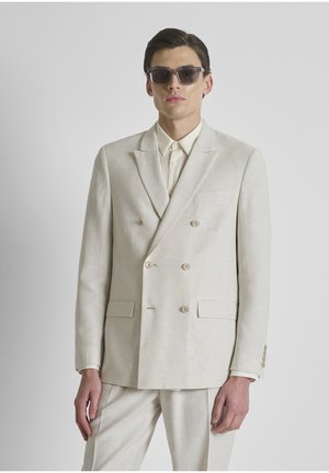 Man draagt een beige dubbelbreasted pak, bijpassende broek, licht shirt en donkere zonnebril, staand tegen een effen lichte achtergrond.