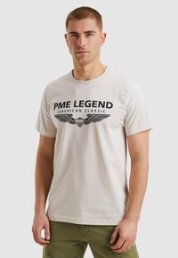Bežna bombažna majica s kratkimi rokavi, ki ima črn tisk z napisom "PME LEGEND AMERICAN CLASSIC" in poudarjenimi krili.