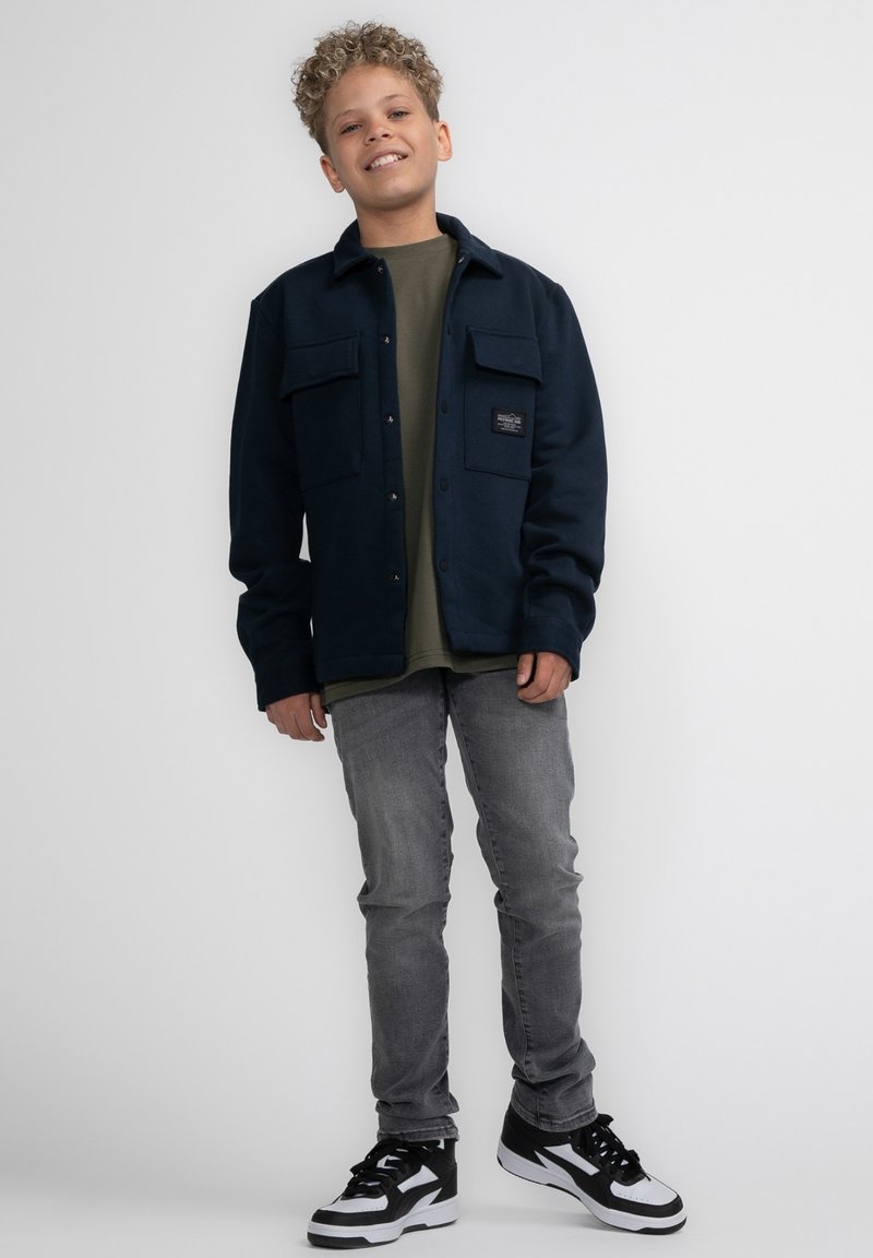 Marineblauwe fleecejas met twee borstzakken, gedragen over een olijfgroen shirt, gecombineerd met grijze jeans en zwart-witte sneakers.