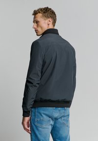 Mannen zwarte bomberjack met een gestructureerd oppervlak, geribbelde kraag en manchetten. Gecombineerd met lichtblauwe denim jeans. Eenvoudig, functioneel ontwerp.