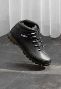 Stivaletto da trekking in pelle nera con colletto imbottito, lacci grigi e suola robusta marrone; presenta accenti metallici e materiali texturizzati.