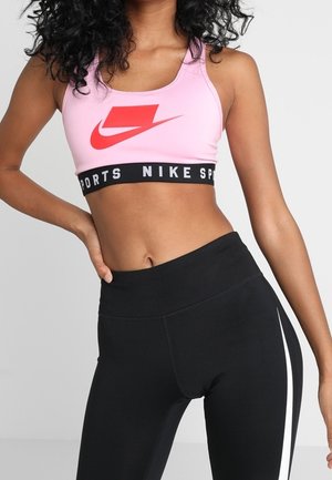 Kobieta w różowym sportowym staniku Nike i czarnych legginsach z białym paskiem, pozująca z jedną ręką na biodrze na jednolitym tle.