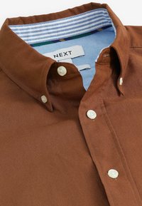 Chemise marron à boutons blancs avec col intérieur rayé, montrant l'étiquette de la marque "NEXT" à l'intérieur du col.