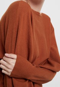 Pullover in Rostfarbe mit entspannter Passform, weitem Ausschnitt und gerippten Bündchen. Das Material wirkt weich und strukturiert, mit minimalen Details.