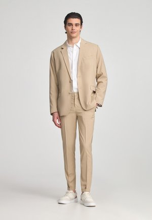 Giacca e pantaloni beige realizzati in tessuto leggero, abbinati a una camicia bianca e sneakers bianche, con un design casual.