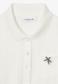 Polo shirt blanc ajusté avec des rayures texturées fines, trois boutons et une poche ornée d'un ruban vert décoratif et d'un emblème de crocodile.