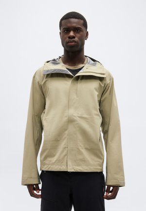 Bărbat purtând o jachetă impermeabilă bej cu glugă și fermoar frontal, pantaloni negri, stând pe un fundal simplu, deschis la culoare.