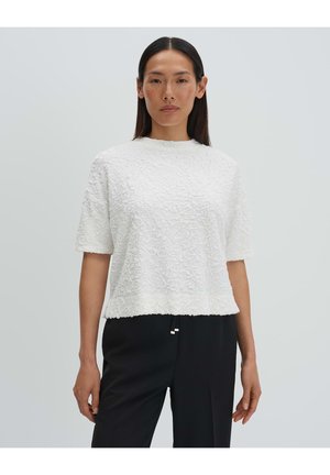 Femme debout face à l'avant, portant un haut à manches courtes texturé blanc et un pantalon noir sur un fond gris uni.