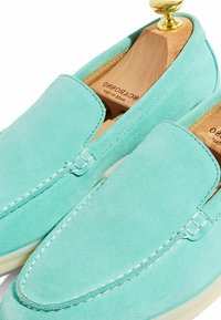 Mocasines de ante en verde claro con costuras blancas en contraste, que presentan una puntera suave y redondeada, y un árbol de zapatos de madera en su interior.