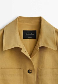 Beige bomullsskjorta med spetsad krage, knapplist fram och två bröstfickor. Har en vävd struktur och en svart varumärkesetikett inuti.