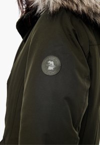 Chaqueta verde oliva con capucha de pelo, que presenta un parche de logotipo metálico redondo en la manga izquierda y una tela suave y texturizada.