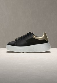 Μαύρο χαμηλό sneaker με λευκή παχιά σόλα και μεταλλική χρυσή λεπτομέρεια στη φτέρνα, παρουσιασμένο πάνω σε ανοιχτή επιφάνεια με ουδέτερο φόντο.