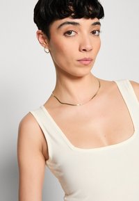 Débardeur couleur crème avec un décolleté carré, en tissu doux et avec des bretelles fines. Un collier en or courbé met en valeur la zone du cou.