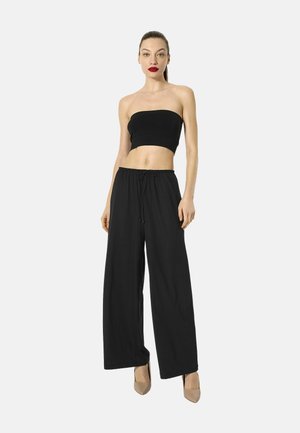 Vrouw staand, gekleed in een zwart strapless crop top, losse zwarte broek met trekkoord in de taille en beige hoge hakken op een witte achtergrond.