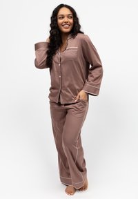 Set di pigiama marrone in tessuto morbido, con top con bottoni e taschino sul petto bordato di bianco, e pantaloni ampi.
