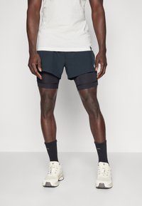 Svarta träningsshorts med ett lager-på-lager-design över tajta kompressionsshorts, kombinerat med vita sneakers och svarta ankelsockor.