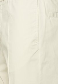 Béžové kalhoty vyrobené z měkké tkaniny, s plochým předním dílem, bočními kapsami a hladkým povrchem s jemnými švy.