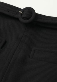 El tejido de sarga negro presenta una cinturilla ancha con un detalle de bucle y un bolsillo rectangular. La textura es suave con sutiles líneas horizontales.