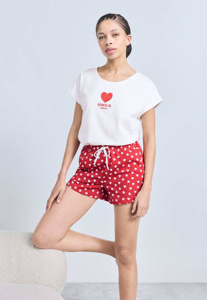 Λευκό μπλουζάκι με κόκκινη καρδιά και κείμενο "AMOUR", συνδυασμένο με κόκκινα σορτς που έχουν λευκά μοτίβα καρδιάς και ελαστική μέση με κορδόνι.