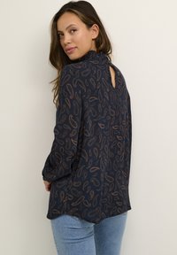 Námornícka modrá blúzka s dlhým rukávom s hnedým paisley vzorom. Má vysoký výstrih a malý otvor na chrbte. Vyrobená z ľahkej látky.