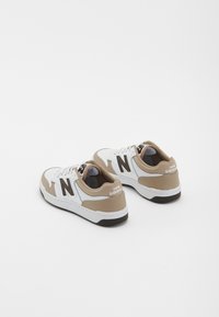 Valkea ja beige New Balance -tennaripari mustilla "N" logoilla sivuilla ja valkoisilla pohjilla yksinkertaisella valkoisella taustalla.