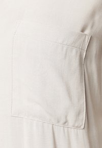 Vaalea beige kangaspaita, jossa on rintatasku, hienovarainen tekstuuri ja näkyvät saumat reunoilla. Yksinkertainen malli ja rento istuvuus.