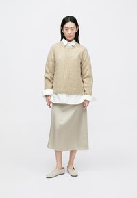 Μπεζ πλεκτό πουλόβερ με υφή, φορεμένο πάνω από λευκό πουκάμισο με γιακά, συνδυασμένο με μπεζ σατέν φούστα και ανοιχτό γκρι παπούτσια χωρίς κορδόνια.