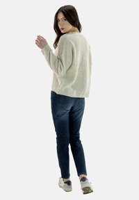 Lichtbeige gebreide trui met een losse pasvorm, gecombineerd met blauwe denim jeans en witte sneakers. De trui heeft een gemêleerd patroon.