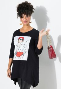 Schwarze Tunika mit einem Punktmuster auf der Vorderseite, die eine rote Handtasche zeigt. Lange Ärmel, lockere Passform und ein abgerundeter Saum.