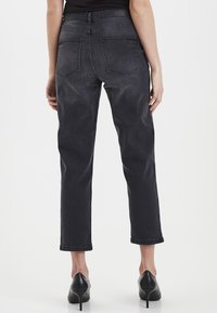 Jean en denim noir avec une jambe droite, taille mi-haute, poches arrière visibles et une texture légèrement décolorée. Portés avec des chaussures à talons noires.