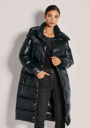 Zwarte pufferjas met glanzende afwerking, oversized kraag, knoopdetails en een doorgestikt patroon; gecombineerd met donkere jeans en een getextureerde top.