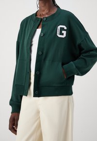 Dunkelgrüner, verkürzter Sweatshirt mit Rundhalsausschnitt, Knopfverschluss, Fronttaschen und einem weißen gestickten Buchstaben "G" als Akzent. Weicher Stoff.