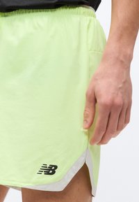 Δεξί χέρι ακουμπισμένο δίπλα σε αθλητικές σορτσάκια νέον κίτρινου χρώματος με μαύρο λογότυπο "NB" και ανοιχτό γκρι περιθώριο, που φοριούνται με μαύρο τοπ.