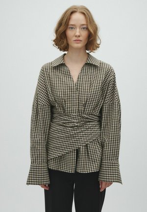 Jeune femme aux cheveux clairs et ondulés porte un chemisier cache-cœur à carreaux avec de larges manches et un pantalon noir sur un fond uni.