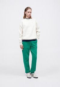 Krēmkrāsas crewneck džemperis, uzvilkts virs tumši zilas blūzes, kopā ar zaļām sporta biksēm un pelēkiem sporta apaviem. Vienkāršs dizains, ikdienišķa piegriezne.