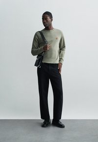 Sweatshirt vert olive avec un petit détail brodé, associé à un pantalon noir à jambes larges et des chaussures oxford noires, rehaussé par un sac bandoulière noir.