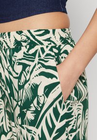 Zelené a slonovinové kalhoty s vzorem, elastickým pasem a bočními kapsami. Tkanina má potisk tropických listů a měkkou strukturu.