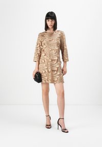 Beige kjole med intrikate, svingende broderier, 3/4 lange ermer og en avslappet passform. Kombinert med svarte strappy hæler og en liten håndveske.