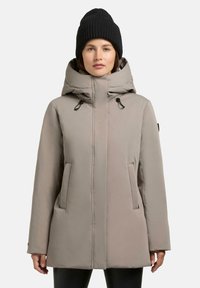 Beige polstret vinterjakke med høj krave, justerbar snorehætte og to forlommer. Båret med en sort strikket beanie.