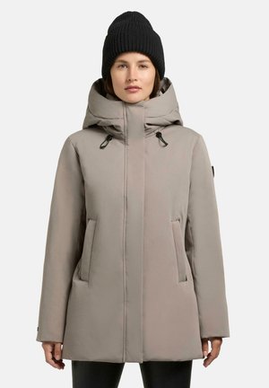 Beige gewatteerde winterjas met een hoge kraag, verstelbare trekkoordcapuchon en twee voorzakken. Draag met een zwarte gebreide beanie.